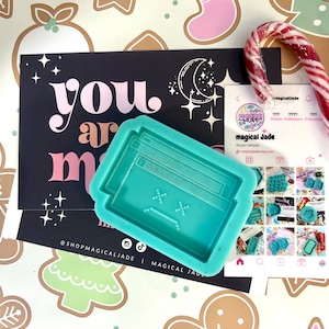 May include: A teal resin mold in the shape of a computer window, with the text "you are magic" on a black card. A candy cane and business card are also present, all on a festive background with gingerbread men and Christmas decorations.