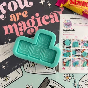 May include: A teal blue silicone mould shaped like a keyboard with the letters W, Q, 2, and A. The mould is on a light blue background with a white daisy and a jar with a bee.