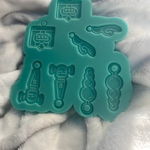 May include: A teal silicone mold with eight different shapes, including a website icon with the text "XXX", a microphone, a beaded necklace, and a pair of wings.