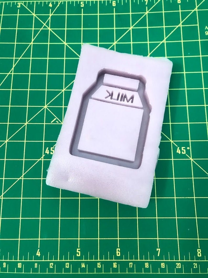 May include: A white silicone mold for making a milk carton shaped cake or cookie. The mold has the word "MILK" embossed on the front.