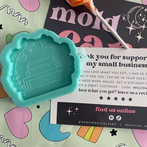 以下が含まれることがあります： 花柄のアクセントが付いた、本を積み重ねた形をしたターコイズブルーのシリコンモールド。モールドは緑とピンクの背景に置かれており、感謝のメッセージカードには「私の小さなビジネスをサポートしてくれてありがとう。あなたが手に入れたものが大好きで、あなたのために作るのはとても楽しかったです。あなたの作品に私をタグ付けしてください。もっとモールドが必要ですか？素晴らしい顧客であるあなたのために割引があります - コード：WELCOME。手に入れたものが気に入りましたか？レビューを残してください。」と書かれています。カードには星評価と「オンラインで私たちを見つける」というテキストとソーシャルメディアのアイコンも表示されています。