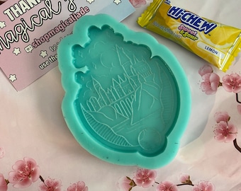 May include: A teal blue silicone mold shaped like a castle with a crescent moon and stars. The mold is on a pink and white background with pink flowers.