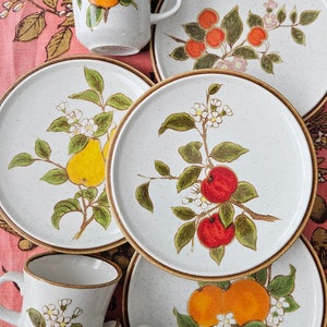 May include: A set of vintage ceramic mugs and plates featuring a floral and fruit design. The mugs and plates have a white background with brown, green, yellow, orange, and red accents. The design includes pears, apples, oranges, and flowers.