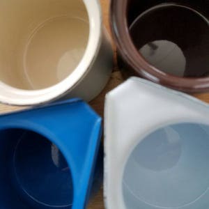 May include: Four ceramic mugs in different colors: white, brown, blue, and light gray. The mugs are all empty and have a smooth, glossy finish.