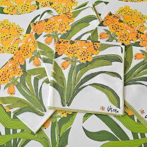 May include: Set of six white napkins with a green and yellow floral design. The napkins feature a stylised flower with yellow petals and green leaves. The napkins are trimmed with a yellow border and have the text "Uska" printed in black on the bottom right corner.