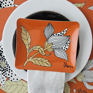 May include: An orange rectangular plate with a floral design, featuring white and black details, rests on a white plate and a black plate. A white napkin is placed beneath the plate. The background is an orange placemat with a matching floral pattern.