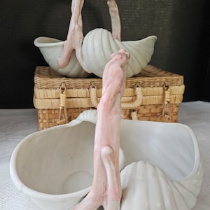 May include: Two white ceramic shell-shaped baskets with pink twig handles. The baskets are sitting on a wicker basket.