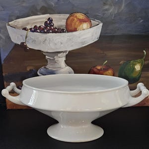 May include: A white ceramic tureen with a wide, shallow bowl, a pedestal base, and two decorative handles on opposite sides. The smooth, glossy finish of the vessel reflects light, highlighting its clean, curved lines and symmetrical design.