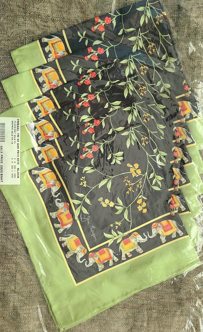 K&ouml;nnte beinhalten: Ein Set aus schwarzen Tischsets und Servietten mit gr&uuml;nem Rand. Die Tischsets und Servietten zeigen ein sich wiederholendes Muster aus Elefanten, Blumen und Ranken. Der Text "Jim Thompson" ist sichtbar.