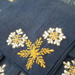 May include: Set of four blue linen napkins with embroidered snowflakes. Two snowflakes are white with gold accents, and two are gold with white accents.