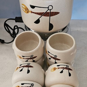May include: Cream-coloured fondue pot with lid and six matching bowls. Each piece features a brown, black, and yellow graphic design. The fondue pot sits on a chrome base with a black power cord.