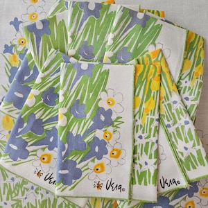 May include: A stack of white linen napkins with a green, blue, and yellow floral pattern. The napkins have a green border and the text "Usagi" is printed on the napkins.