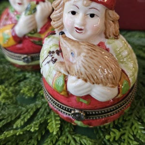 May include: Two decorative ceramic trinket boxes. Each box features a figure with a red hat and a detailed face. One figure holds a small bird, the other a hedgehog. The boxes are red, green, and white, with gold accents.