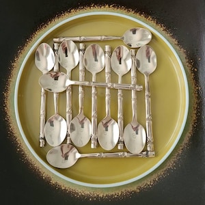 May include: A set of 12 silver teaspoons with bamboo-style handles arranged on a yellow plate with a black and gold rim.