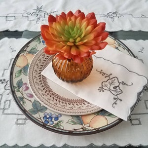 Op de afbeelding: Een witte tafelsetting met een bord met een bloemenpatroon, een witte servet met een geborduurd ontwerp en een glazen vaas met een vetplant. De vetplant is oranje en groen.