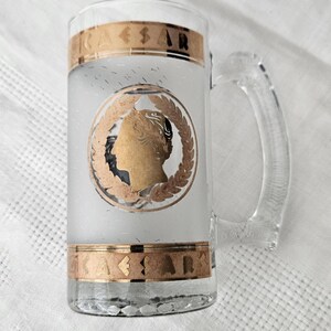 May include: Clear glass beer mug with a handle, featuring a gold-colored band at the top and bottom with the word "CAESAR". The mug has a frosted center with a gold profile of a person in a laurel wreath.
