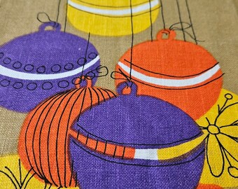 May include: A brown fabric kitchen towel with a colorful design of hanging ornaments. The ornaments are purple, orange, yellow, and red with white accents. The towel has the text "Velva" and a red circle with the text "VIVA" and "kitchen originals" printed on it.