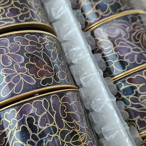 May include: A set of decorative, cylindrical containers with a floral design. The containers are primarily dark blue with purple and gold accents. The floral pattern is outlined in gold. The containers are stacked and wrapped in plastic.