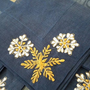 May include: Set of four blue linen napkins with embroidered snowflakes. Two snowflakes are white with gold accents, and two are gold with white accents.