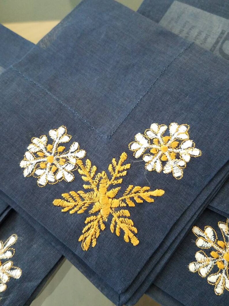 May include: Set of four blue linen napkins with embroidered snowflakes. Two snowflakes are white with gold accents, and two are gold with white accents.