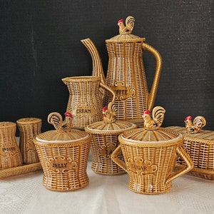 May include: A set of brown ceramic kitchenware with a woven wicker pattern and rooster accents. The set includes a coffee pot, creamer, sugar bowl, jelly jar, and two pepper shakers. Each piece is labelled with its contents: "Coffee", "Cream", "Sugar", "Jelly", and "Pepper".