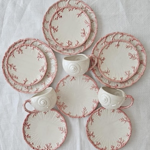 May include: Set of six white ceramic teacups and saucers with pink coral designs. The cups and saucers have a raised rim and a scalloped edge.