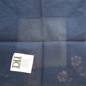 May include: A dark blue square fabric napkin with a white embroidered design. The napkin has a label that reads "DII Home Fashions 100% Cotton Made in India Hand Wash Cold Line Dry Low Iron"