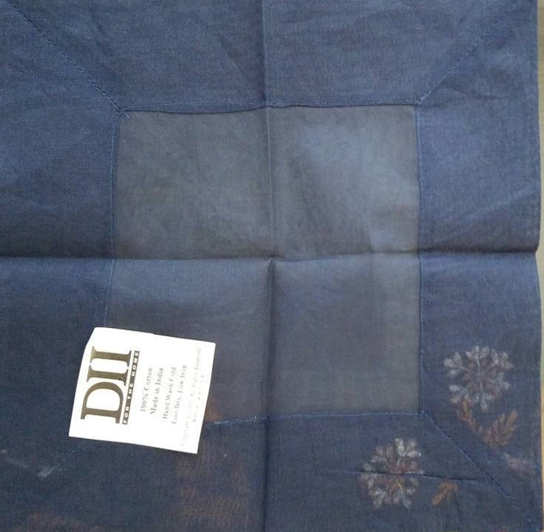 May include: A dark blue square fabric napkin with a white embroidered design. The napkin has a label that reads "DII Home Fashions 100% Cotton Made in India Hand Wash Cold Line Dry Low Iron"