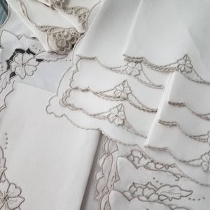 Puede incluir: Un juego de servilletas de lino blanco con delicados diseños florales bordados. Las servilletas tienen bordes festoneados y están dobladas cuidadosamente.