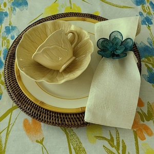 May include: A white napkin with a teal blue floral napkin ring sits on a brown wicker charger plate. A white plate with a yellow leaf-shaped bowl and a cream-colored teacup sits on the charger plate. The background is a white tablecloth with a green and orange floral pattern.