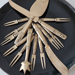 May include: A set of silver-toned cutlery on a black plate. The set includes a butter knife, a serving spoon, and several small forks.