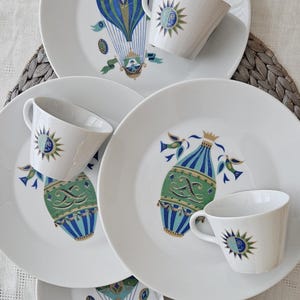 May include: White ceramic plates and cups with a hot air balloon and sunburst design in blue, green, and gold. The plates and cups are arranged on a neutral-colored surface, creating a cohesive tableware set.