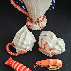 Peut inclure: Une théière en céramique blanche et deux tasses en forme de coquillages avec des accents de corail. Deux figurines de crevettes rouges et blanches sont également incluses.