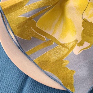 May include: A yellow and blue floral patterned napkin with a gold napkin ring. The napkin is folded and placed on a white plate.