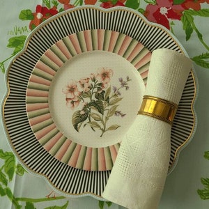 May include: A white napkin with a gold napkin ring sits on a black and white striped plate with a pink and white floral design. The plate is on a light blue tablecloth with a floral pattern.