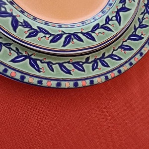 May include: Two ceramic plates with a green, blue and orange floral design. The plates are stacked on top of each other and are sitting on a red surface.