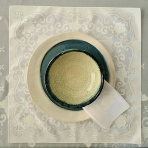 May include: A white ceramic bowl with a crackled glaze sits inside a teal blue bowl, which is inside a white plate. The bowls and plate are on a white lace tablecloth with a floral pattern.