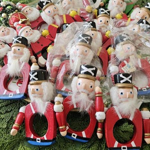 May include: A collection of wooden Christmas ornaments featuring Santa Claus and toy soldier designs. The ornaments are primarily red, white, and black, with some in plastic packaging. The background is a bed of green foliage.
