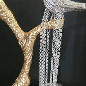 May include: A silver chain necklace displayed on a gold decorative stand shaped like a bird. The chain is made of small, linked circles.