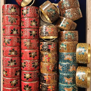 May include: A collection of decorative napkin rings in various colors. The rings feature floral designs in red, brown, blue, and gold, with gold-colored rims. The rings are arranged in stacks and rows, showcasing their intricate patterns.