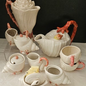 May include: A seashell-themed tea set with a teapot, creamer, sugar bowl, and cups. The set is made of white ceramic with red and pink accents. The teapot has a coral-like handle and a seashell lid.