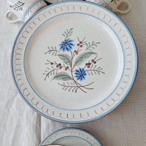 May include: A ceramic dinnerware set featuring a speckled off-white base with blue floral motifs, brown stems, and light blue rim accents. The collection includes a large dinner plate and several mugs, all decorated with a consistent hand-painted pattern.