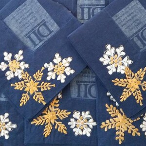 May include: A stack of blue fabric napkins with white and gold embroidered snowflakes. The napkins have the letters "DI" printed on them.