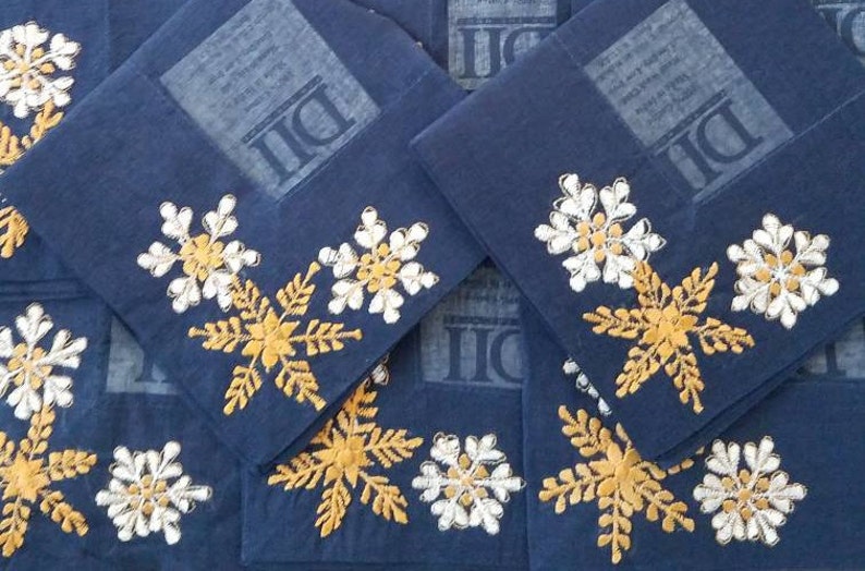 May include: A stack of blue fabric napkins with white and gold embroidered snowflakes. The napkins have the letters "DI" printed on them.