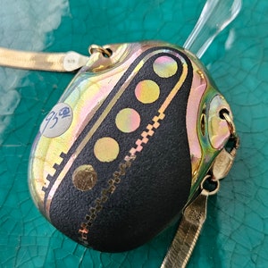 Op de afbeelding: Een gouden ketting met een zwarte en gouden hanger. De hanger is een ronde, iriserende glazen hanger met een geometrisch ontwerp. De hanger heeft een kleine, doorzichtige glazen stop bovenop.