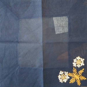 May include: A dark blue fabric square with a sheer panel in the center. The panel has a smaller, darker blue fabric square sewn into the upper right corner. The fabric square has gold and white embroidered flowers in the lower right corner.
