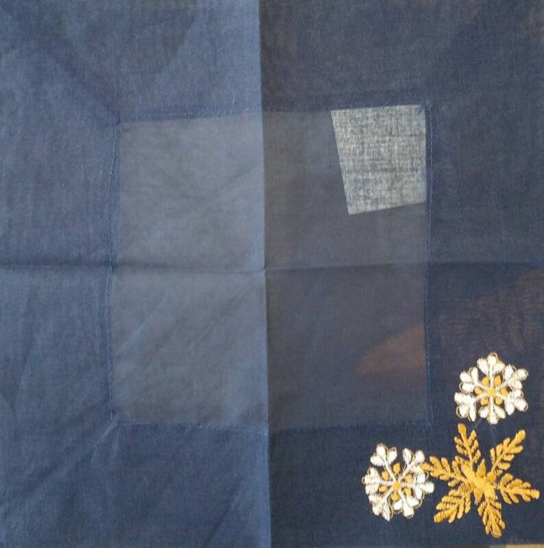 May include: A dark blue fabric square with a sheer panel in the center. The panel has a smaller, darker blue fabric square sewn into the upper right corner. The fabric square has gold and white embroidered flowers in the lower right corner.