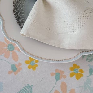 May include: A white linen napkin with a dark blue beaded napkin ring on a white plate with a scalloped edge. The plate is on a white tablecloth with a floral pattern.