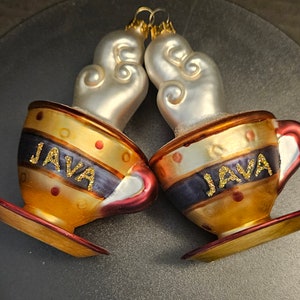 May include: Two gold and white ceramic coffee mugs with red rims and a saucer. The mugs have a black stripe with the word "JAVA" in gold glitter. The mugs are topped with white ceramic steam.