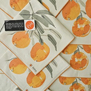 May include: Off-white linen napkins featuring a repeating pattern of oranges and green leaves. Each napkin has an orange border. The design includes the text "Vera" and a small orange logo. The napkins are folded.